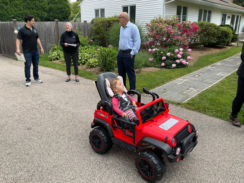 Bear, a young girl wearing a pink shirt, is riding in her specially adapted red motorized Jeep in the driveway of the family home.