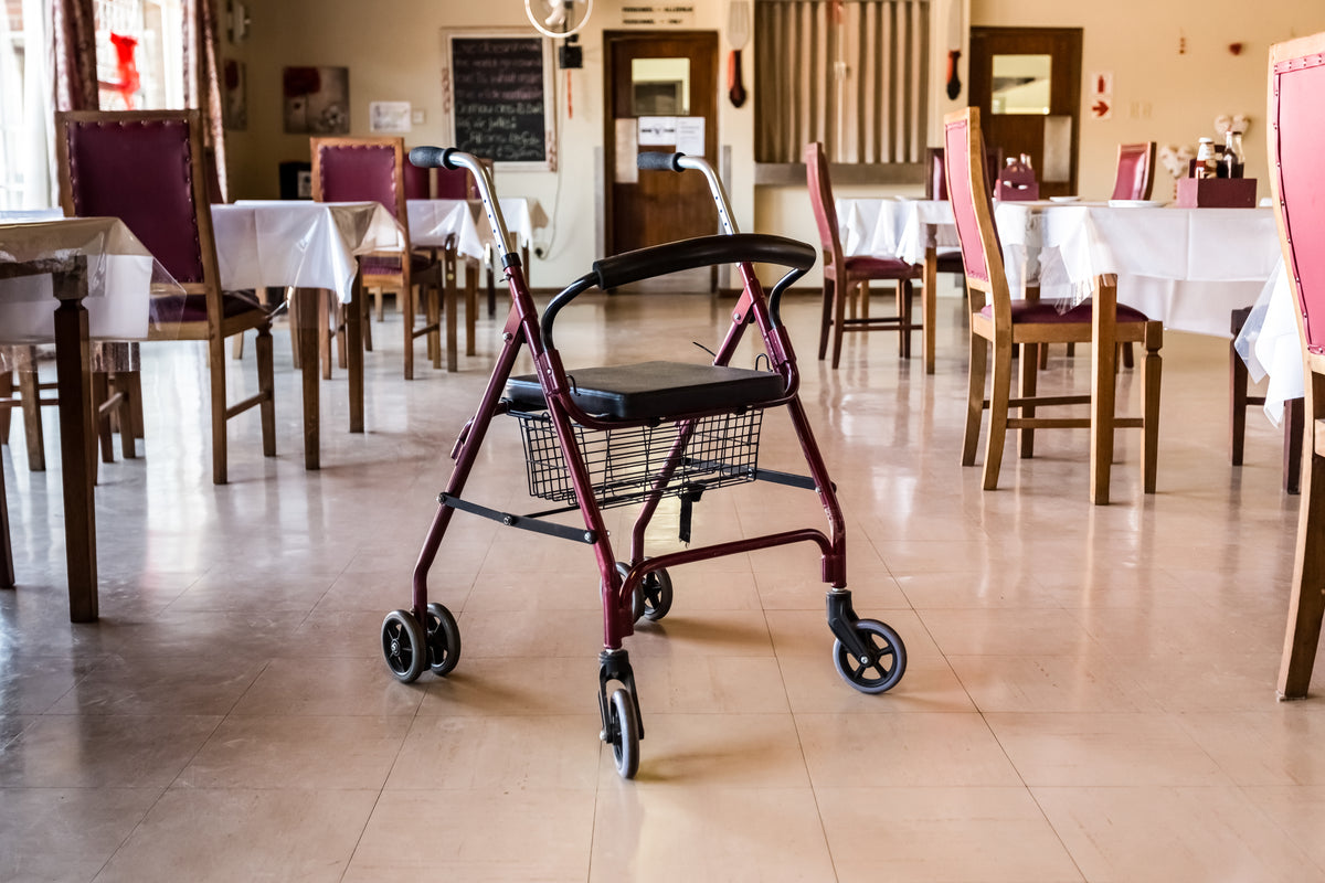 a red rollator walker with a seat and basket is viewed at an angle in a beige room filled with rows of chairs and tables