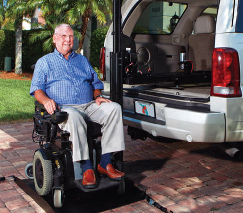 an elderly man in a blue shirt sits in a power wheelchair that is being parked on a hybrid vehicle lift