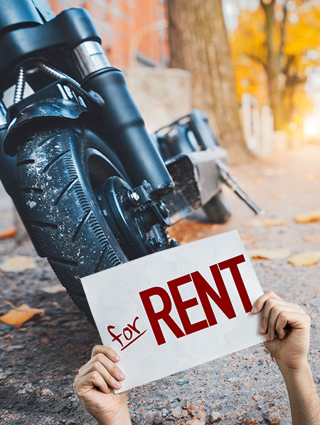 a pair of hands holds up a sign reading 'for RENT' in front of a motorcycle tire