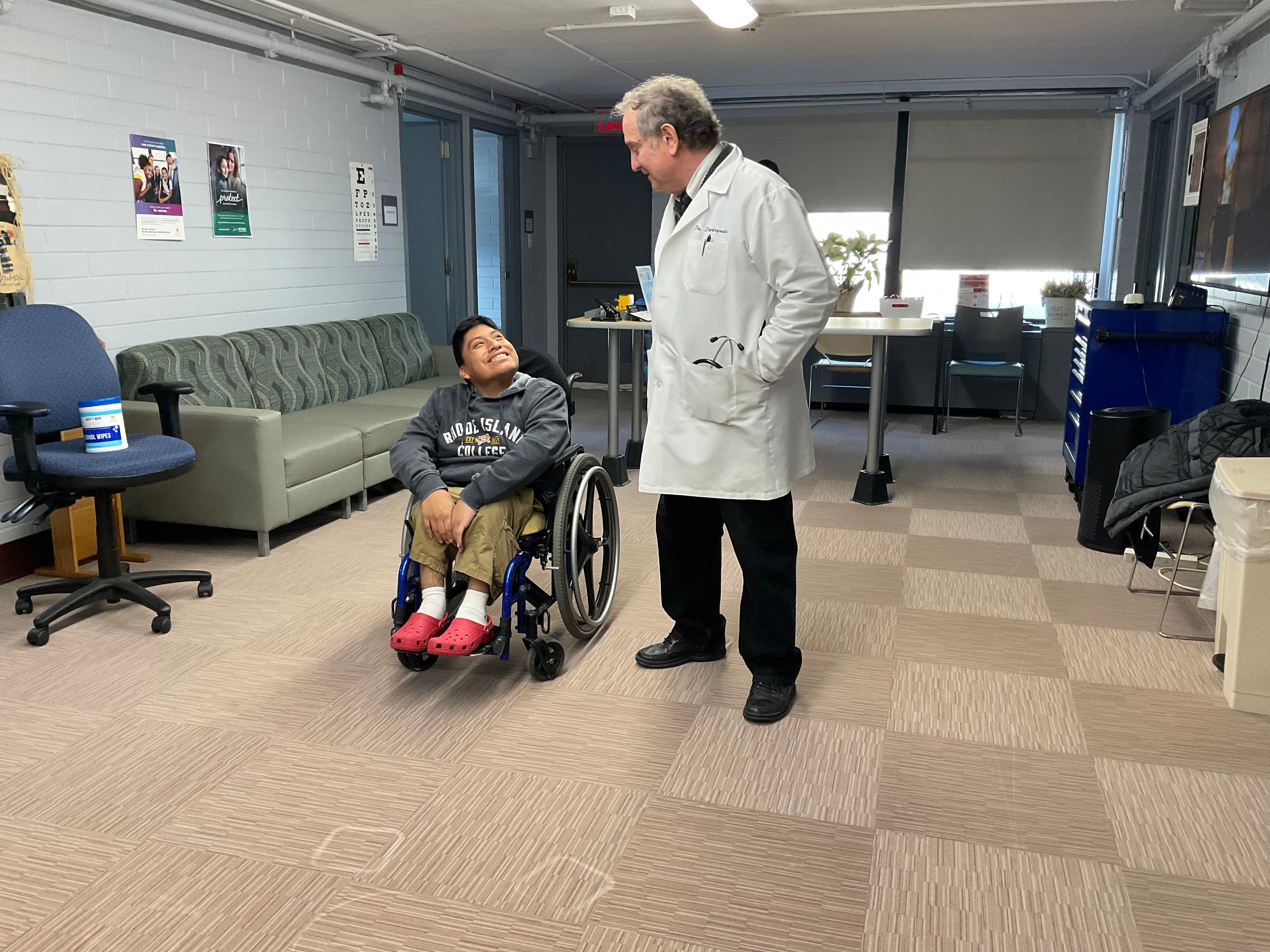 A medical staff member in a lab coat looks at a young man in a wheelchair, the young man looking up at him and smiling 