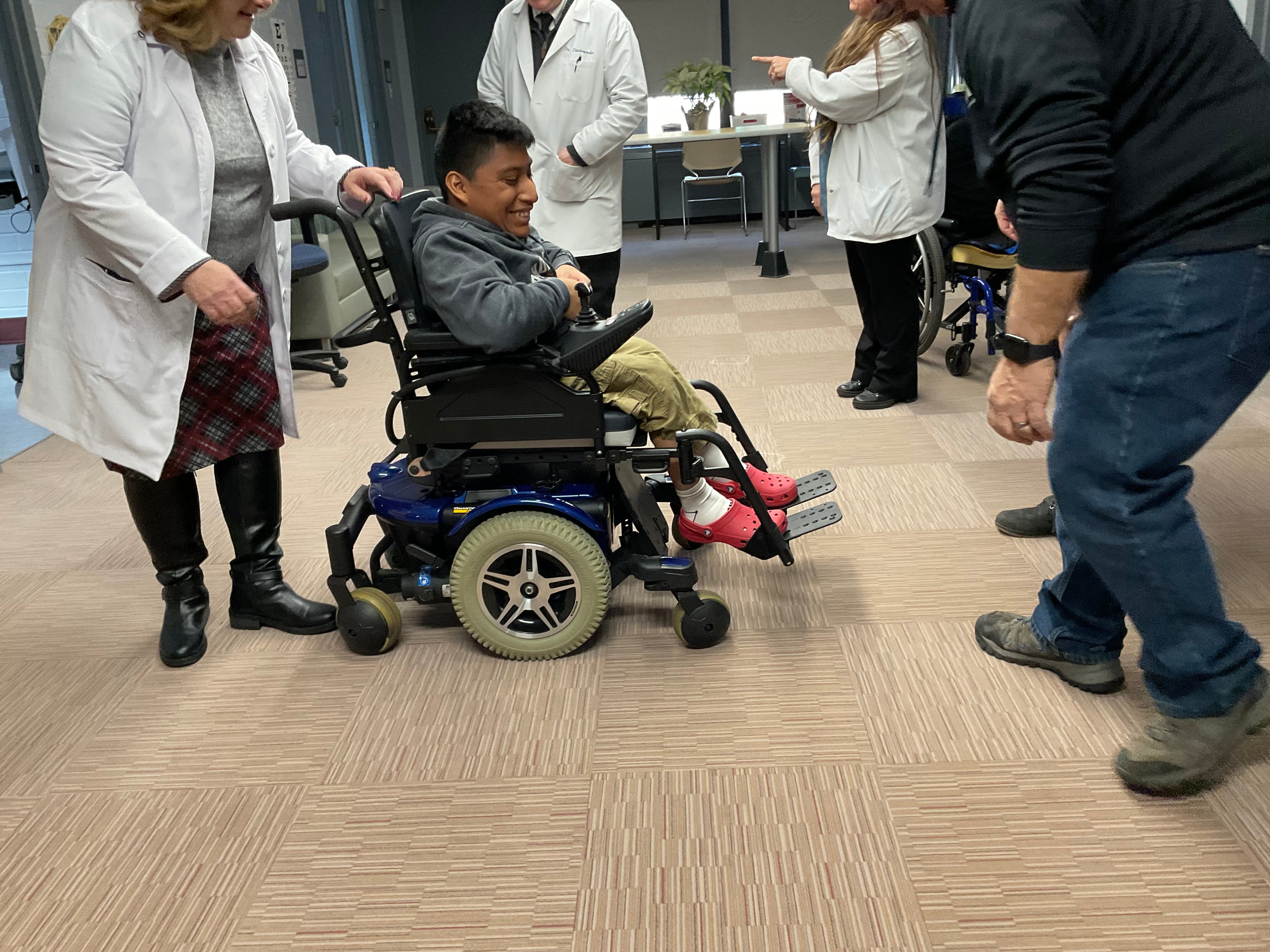 Christian, surrounded by Browne Hall doctors and a mechanic, smiling and looking excited for his new power chair