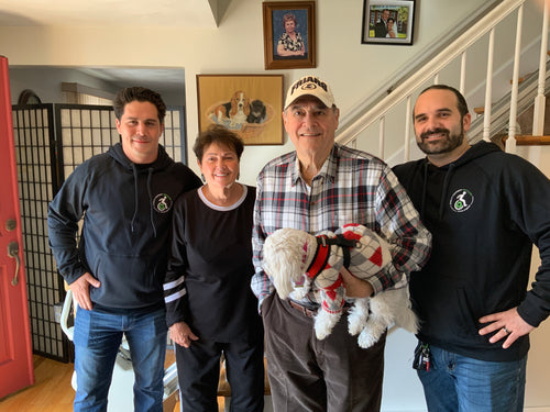 (From left to right) John, Kathi, Mike, (and their white dog), and General Manager Andrew pose for a photo in front of the stairlift in their home. The stairs are visible in the background and a number of framed photos hang on the white wall behind them.