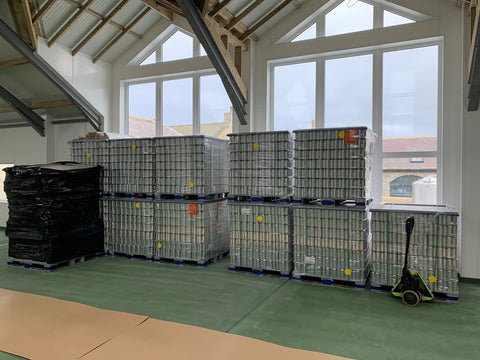 Image shows a series of pallets of beer cans, standing in a brewery ready to have labels applied. There is a large set of windows in the background.