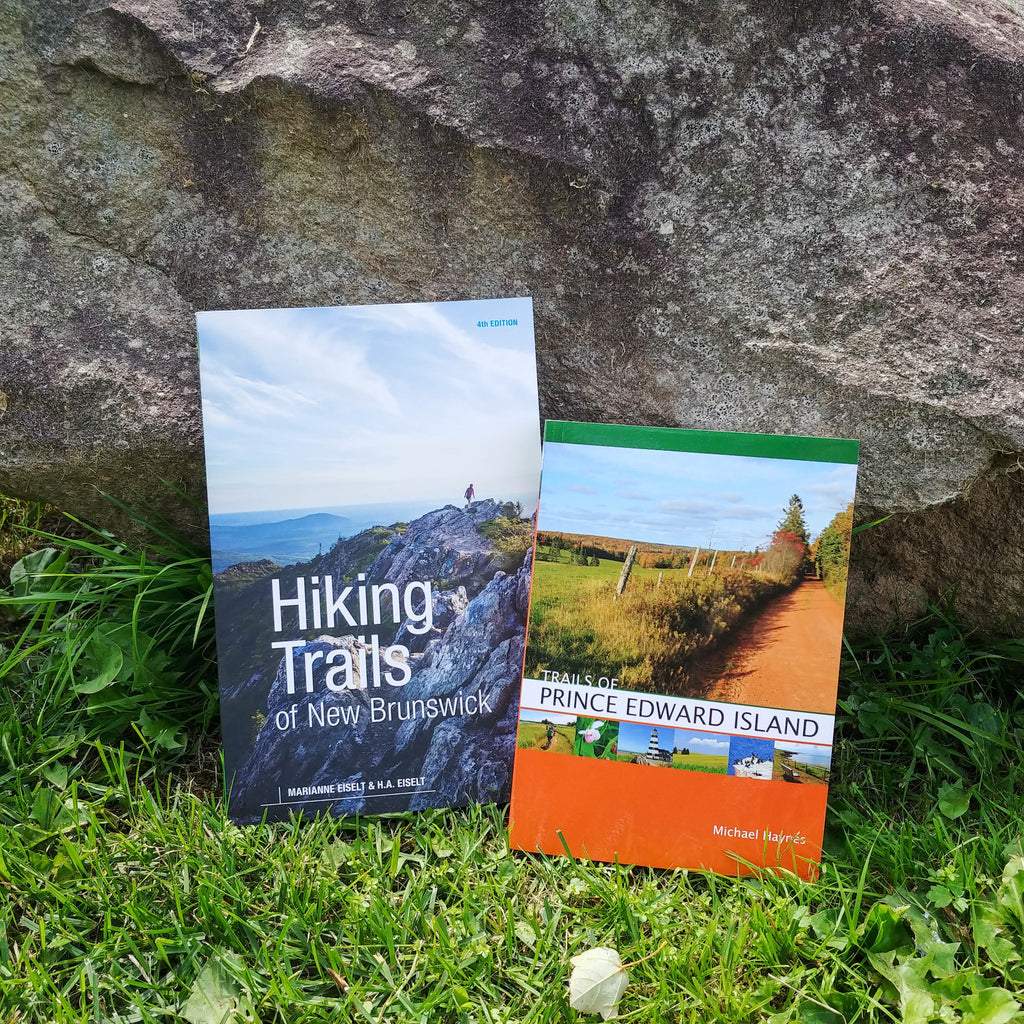 Photo of two books standing in grass, propped up against a boulder. The two books are Hiking Trails of New Brunswick, 4th Edition and Trails of Prince Edward Island.