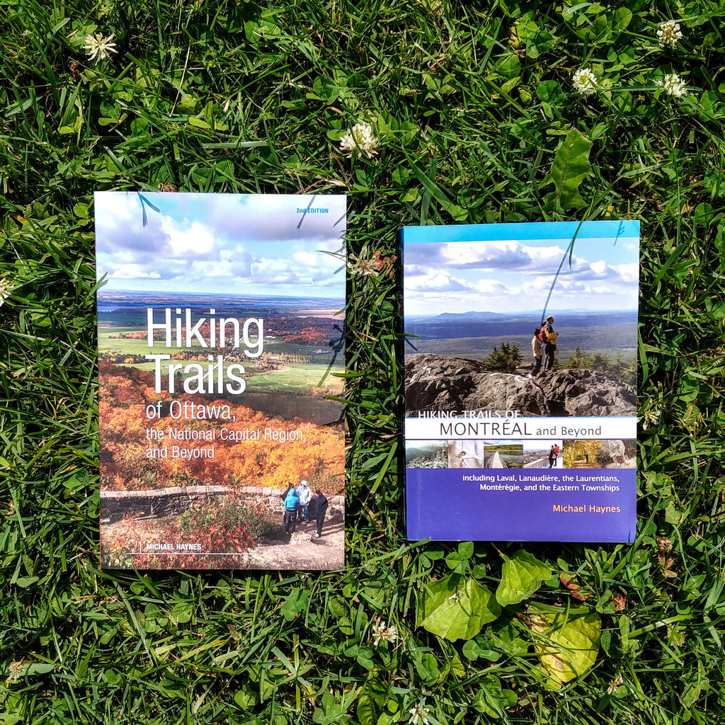 Photo of two books lying in lush green grass. The books are Hiking Trails of Ottawa, The National Capital Region, and Beyond, 2nd Edition and Hiking Trails of Montreal and Beyond.