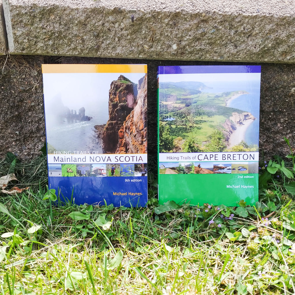 Photo of two books standing in grass, propped up against a rough, stone wall. The two books are Hiking Trails of Mainland Nova Scotia and Hiking Trails of Cape Breton.