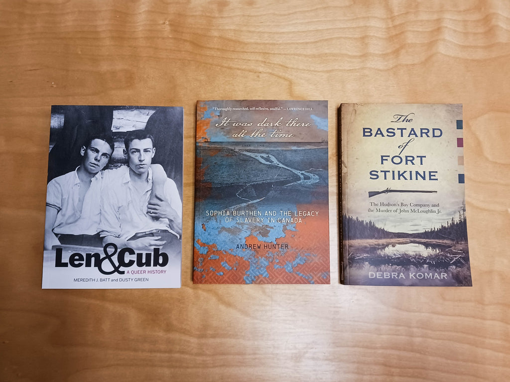 Three books sit on a wooden table, Len & Cub, It Was Dark There All the Time, and The Bastard of Fort Stikine.
