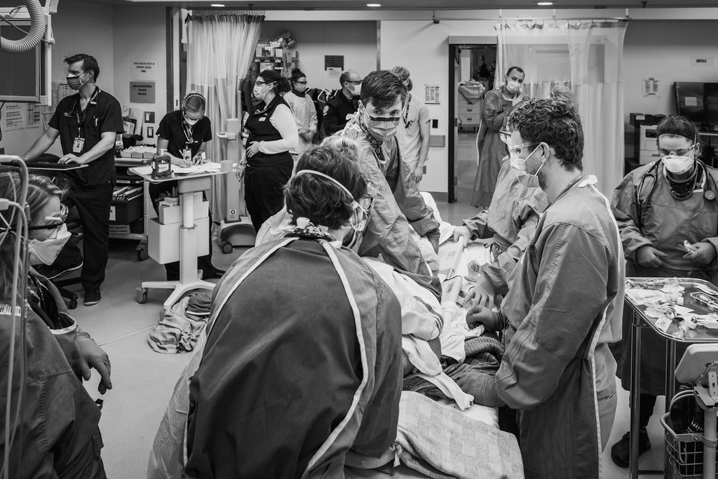 A hospital ER is bustling with activity. In the center of the chaos, several doctors and nurses attend to a patient on a gurney.