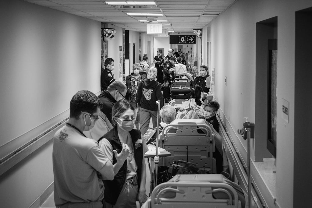 A narrow hospital hallway is packed tight with hospital personnel and visitors. Along the right wall is a long line of patients on gurneys.