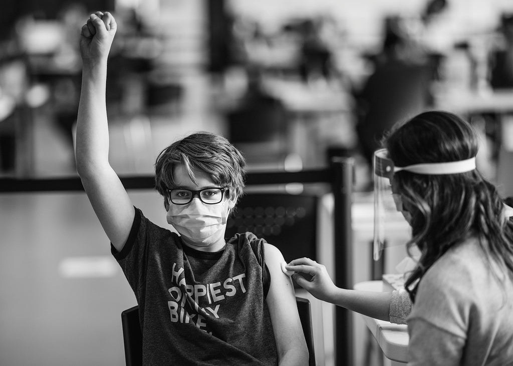 A child, perhaps middle-school aged, holds their left fist in the air as a hospital attendant swabs their upper right arm, preparing for an injection.