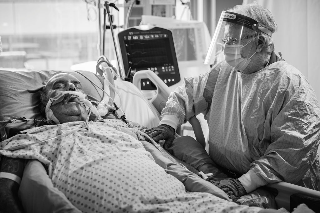 An elderly man on a ventilator lies in a hospital bed, eyes closed, in a hospital gown. An elderly woman in full PPE gear stands to the right of the bed, holding his arm.