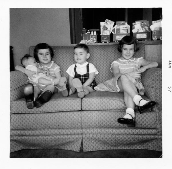 A black and white photo of three children sitting on a couch. One of the children is holding a baby.