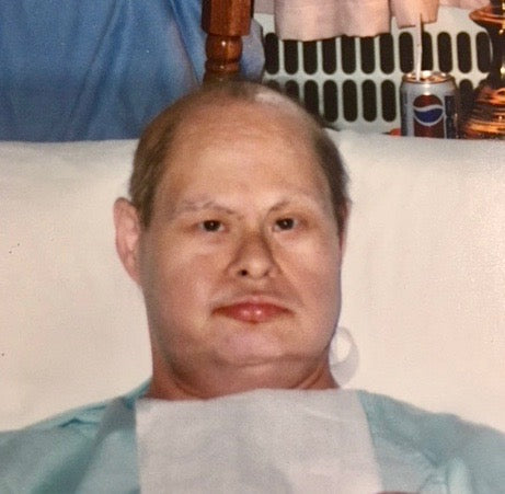 A photo of Bill, from the chest up. Bill appears to be sitting in bed with a pillow behind him, lips upturned in a slight smile.