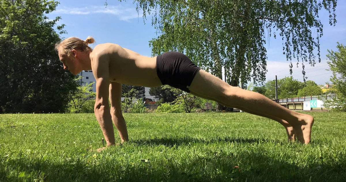 A blond man wearing black trunks holds a plank pose in a grassy park.
