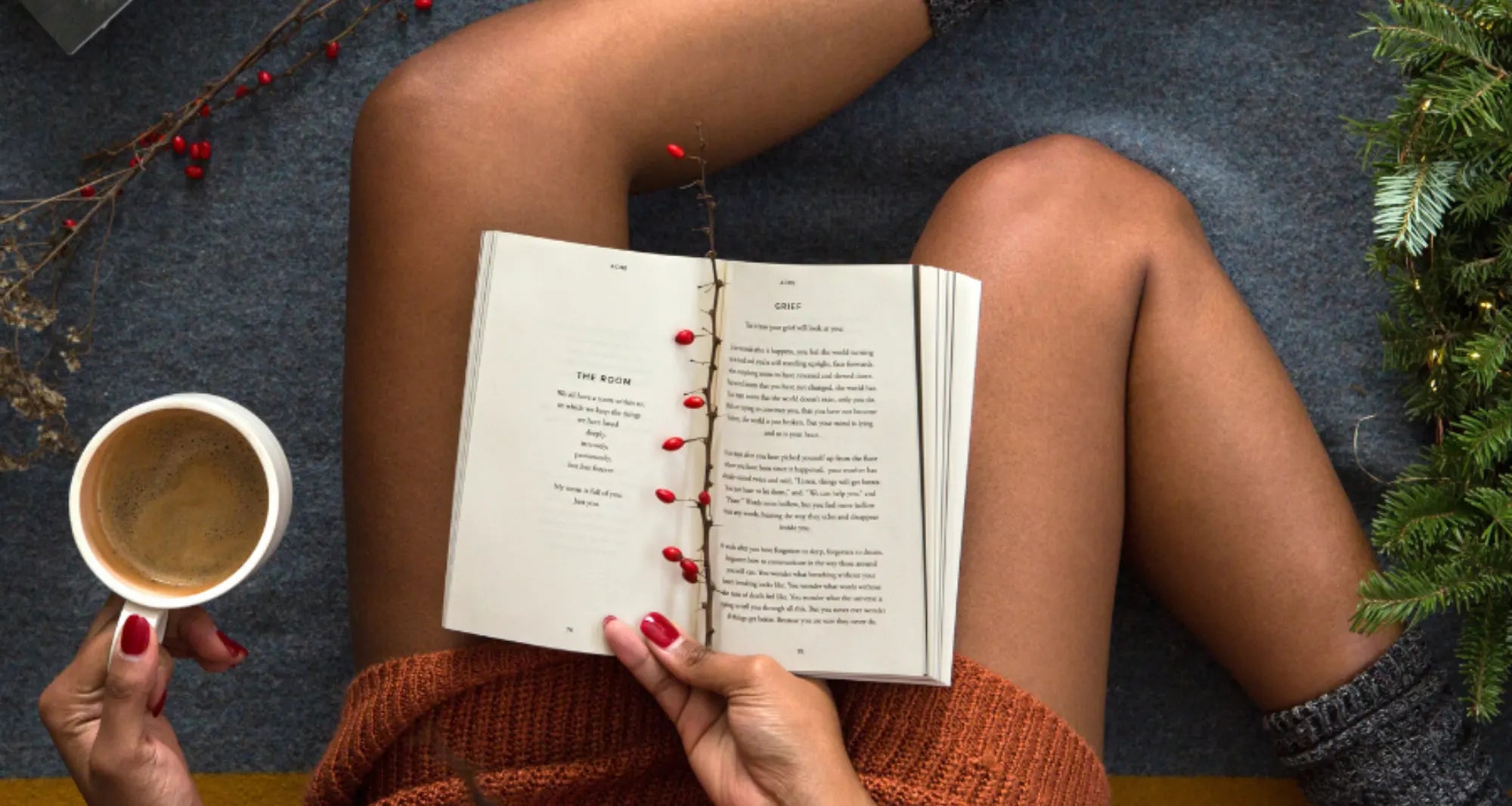 A woman wearing an orange sweater and gray socks reads a book and holds a mug while sitting on a gray blanket surrounded by books, a wreath, and dry flowers.  
