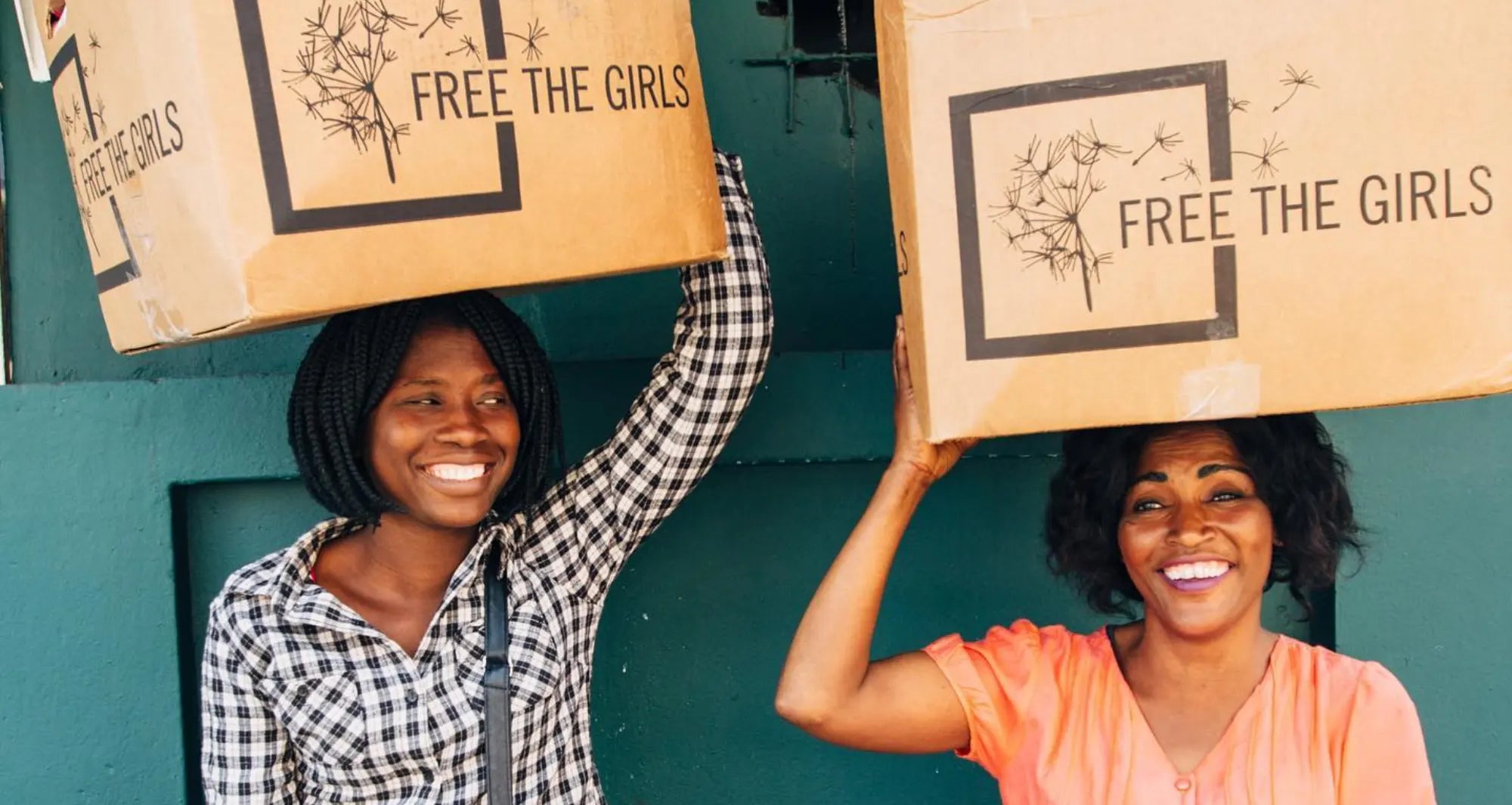 Two women standing side by side against a green wall, each holding a cardboard box with a “free the girls” logo over her head