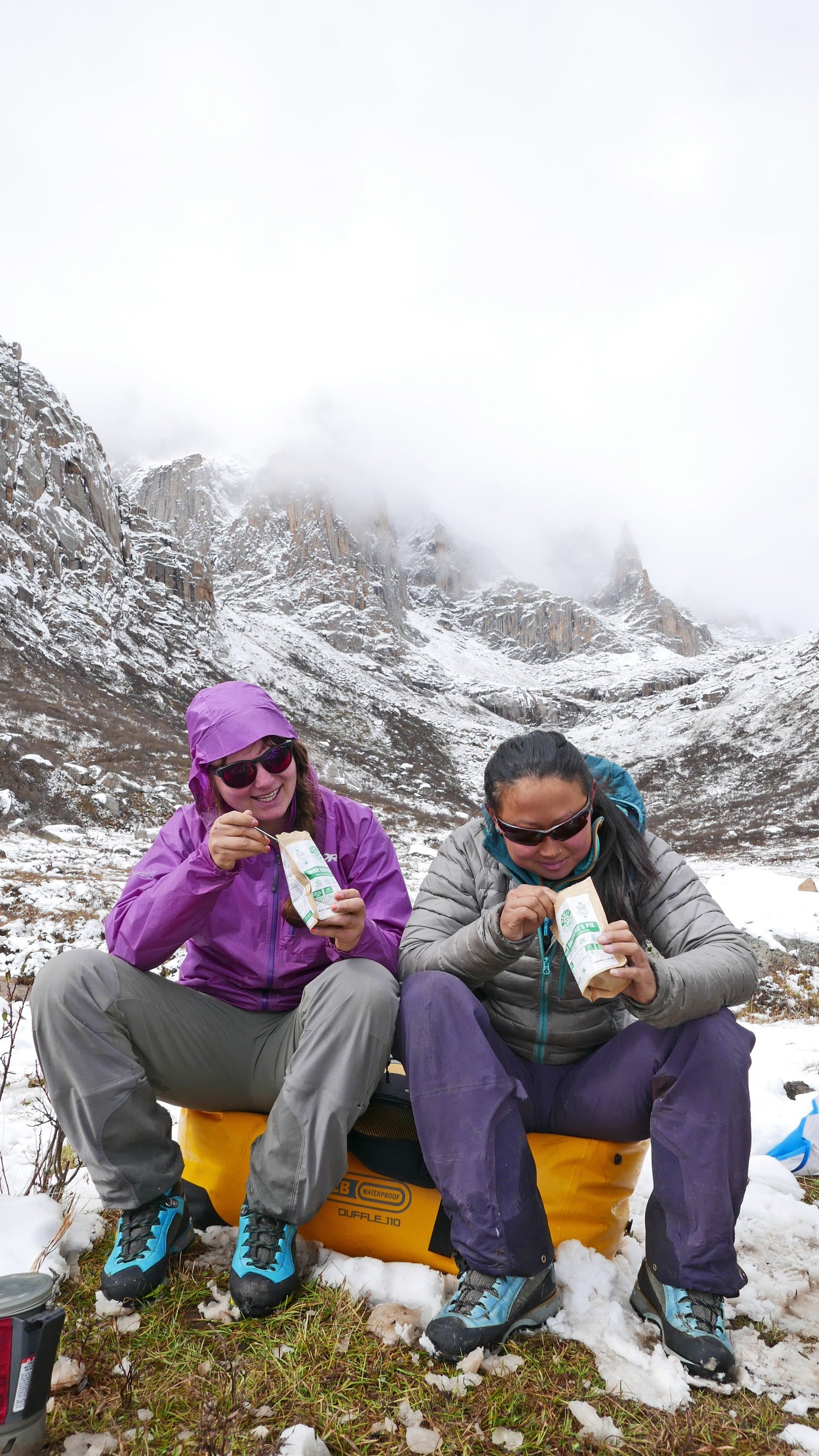 Two people enjoying Nomad Nutrition, plant-based dehydrated meals optimized for nutrients, outdoors in the winter. Foggy snow-capped mountains visible in the background. 