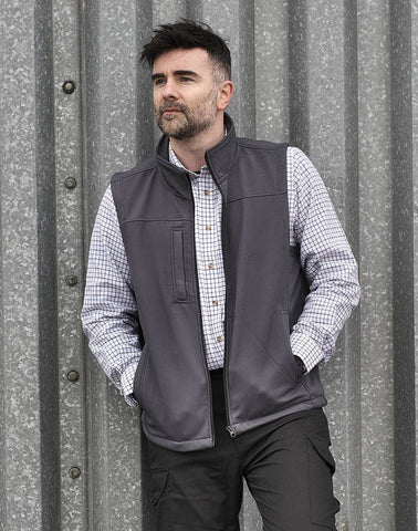 Man wearing tattersall shirt with fleece gilet and trousers against a wall background