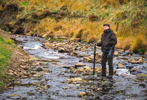 Man wearing Jack Pyke clothing standing on the banks of a river holding a gun for shooting