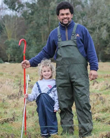 Farmer and child wearing waterproof overalls standing together in a field, smiling and holding up farm equipment