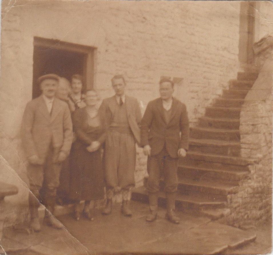 Black and white photo of a family of six people standing at the door step of a stone cottage.