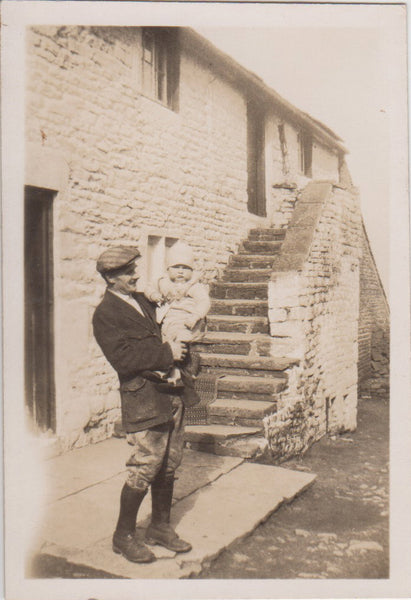 Black and white photo of a farmer holding a baby in front of a stone cottage