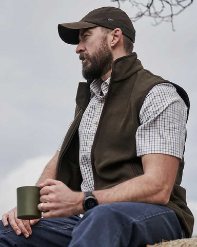 Man wearing baseball cap, jeans, fleece gilet and tattersall shirt sitting down outdoors and holding a mug