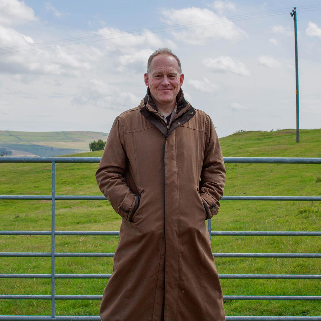 Man in a brown long waterproof coat standing in front of a farm gate