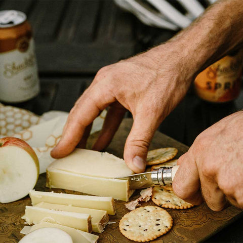 Person using Opinel pocket knife to slice through cheese on a wooden cutting board