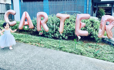 Toddler girl wearing a dress outside in front of birthday balloons that spell "Carter" during drive-by first birthday party