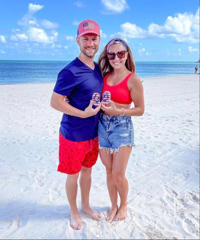 A couple poses on the beach with red white and blue baby flip-flops in a 4th of July pregnancy announcement photo
