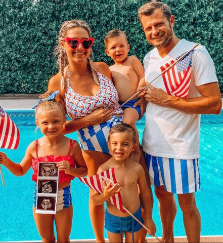 A family poses by the pool in red, white, and blue swimsuits for a 4th of July pregnancy announcement photo