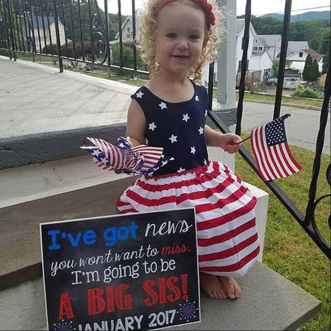A toddler girl poses in a patriotic dress next to a chalkboard in a 4th of July pregnancy announcement