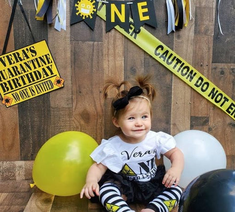 Baby girl sitting in front of a yellow "Vera's Quarantine Birthday" sign and yellow, white, and black balloons