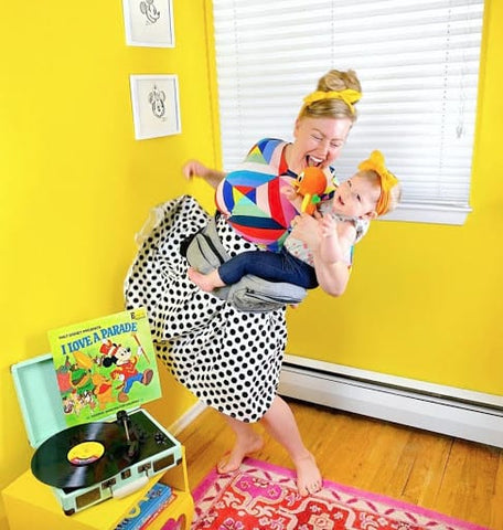 Mother holding 1-year-old baby as she dances to a Mickey Mouse record in a bright yellow room