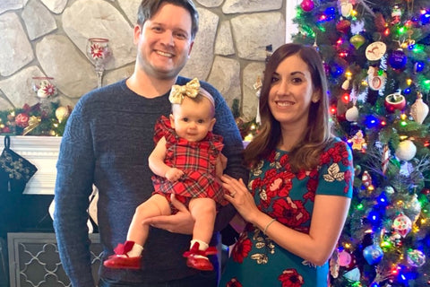 Father, mother, and baby girl standing by a Christmas tree in holiday attire.