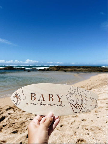 A mini wooden surfboard that says "baby on board" is used in a beach pregnancy announcement
