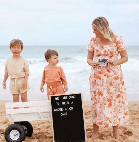 A pregnant mom poses with her two kids and a cart and a sign that says "we're going to need a bigger cart" in a beach pregnancy announcement