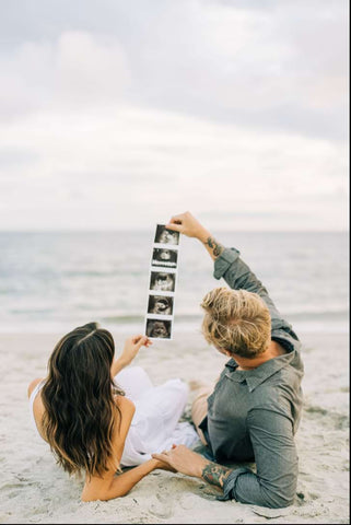 A couple lays on the beach and holds up their ultrasound photos in a beach pregnancy announcement