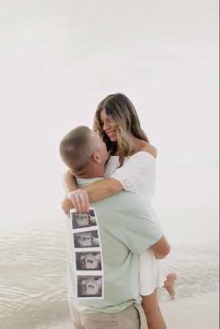 A couple poses with their ultrasound photos in a beach pregnancy announcement