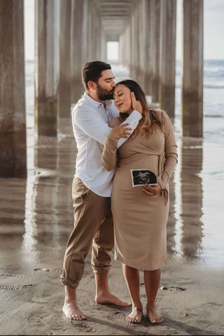 A couple poses with an ultrasound photo under a pier in a beach pregnancy announcement