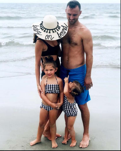 A mom wears a straw hat that says "baby on board" and poses with her family in a beach pregnancy announcement