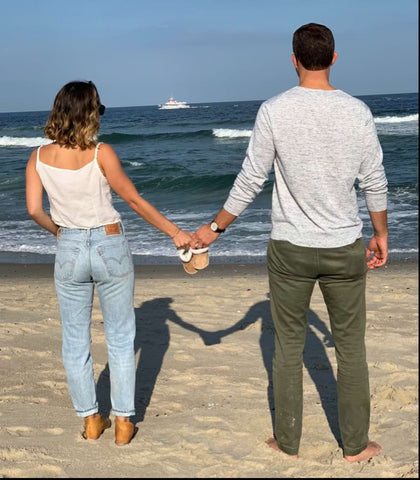 A couple poses with baby shoes on the beach to announce a pregnancy