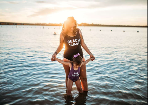 A mom and daughter pose in swimsuits that say "Beach Bump" and "Big Sis" in a beach pregnancy announcement