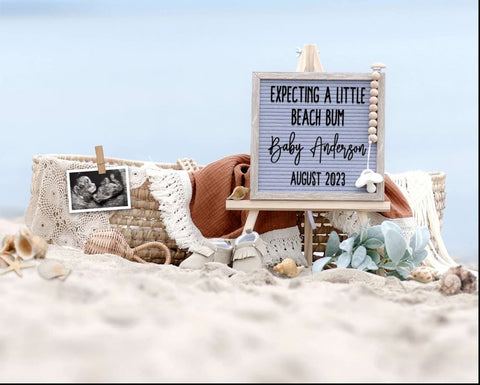 A letterboard and baby items arranged on the sand for a beach pregnancy announcement