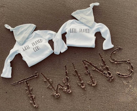 Baby outfits that say "Well Played, God" displayed near the word "Twins" written in sand