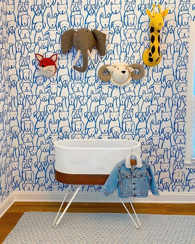 A SNOO bassinet in front of nursery wallpaper printed with a blue hand-drawn-looking pattern of dogs. Above the SNOO there are a plush felt giraffe, elephant, ram, and fox on the wall.