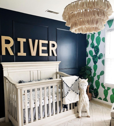 A nursery with a dark blue painted accent wall and the word RIVER printed in gold behind the crib. The adjacent wall has green cactus-print wallpaper.