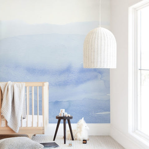A blue baby nursery with a light blue water-color mural on the wall behind the crib.